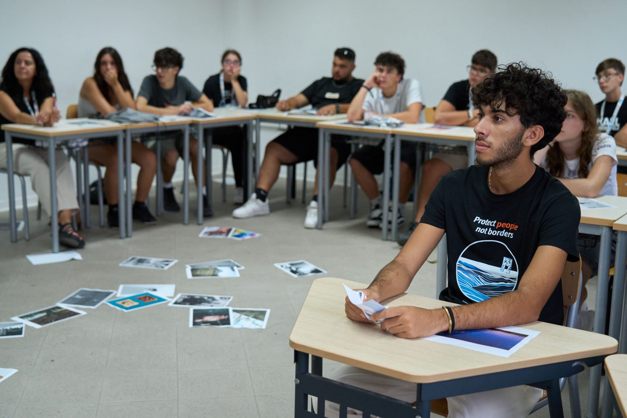 Gruppo di persone in un laboratorio partecipa a un’attività con foto sparse sul tavolo, maglietta con scritto "Protect people not borders".