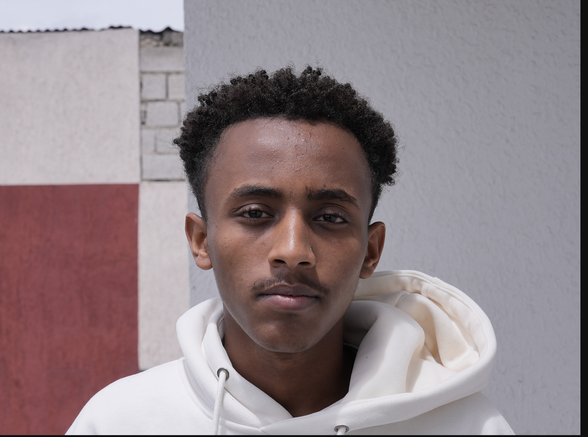 Giovane con hoodie bianco, espressione seria, contro muro grigio e rosso, inquadratura frontale.