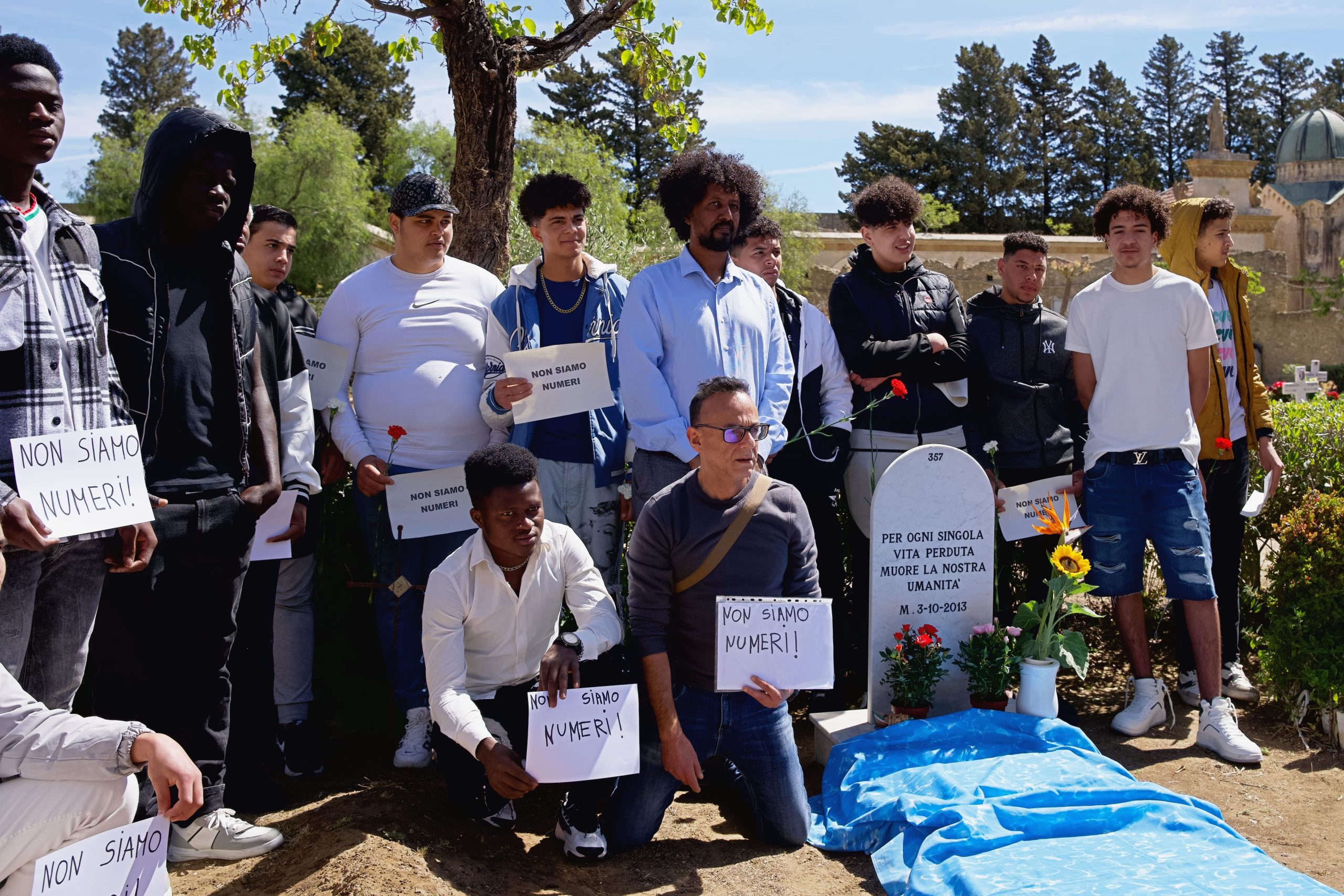 Persone in gruppo davanti a una tomba con cartelli che recitano "Non siamo numeri!", in un cimitero con fiori e bandiere.