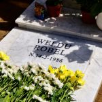 Lapide con il nome "Weldu Robel" e data "N.14-2-1986", circondata da fiori bianchi e gialli in un cimitero.