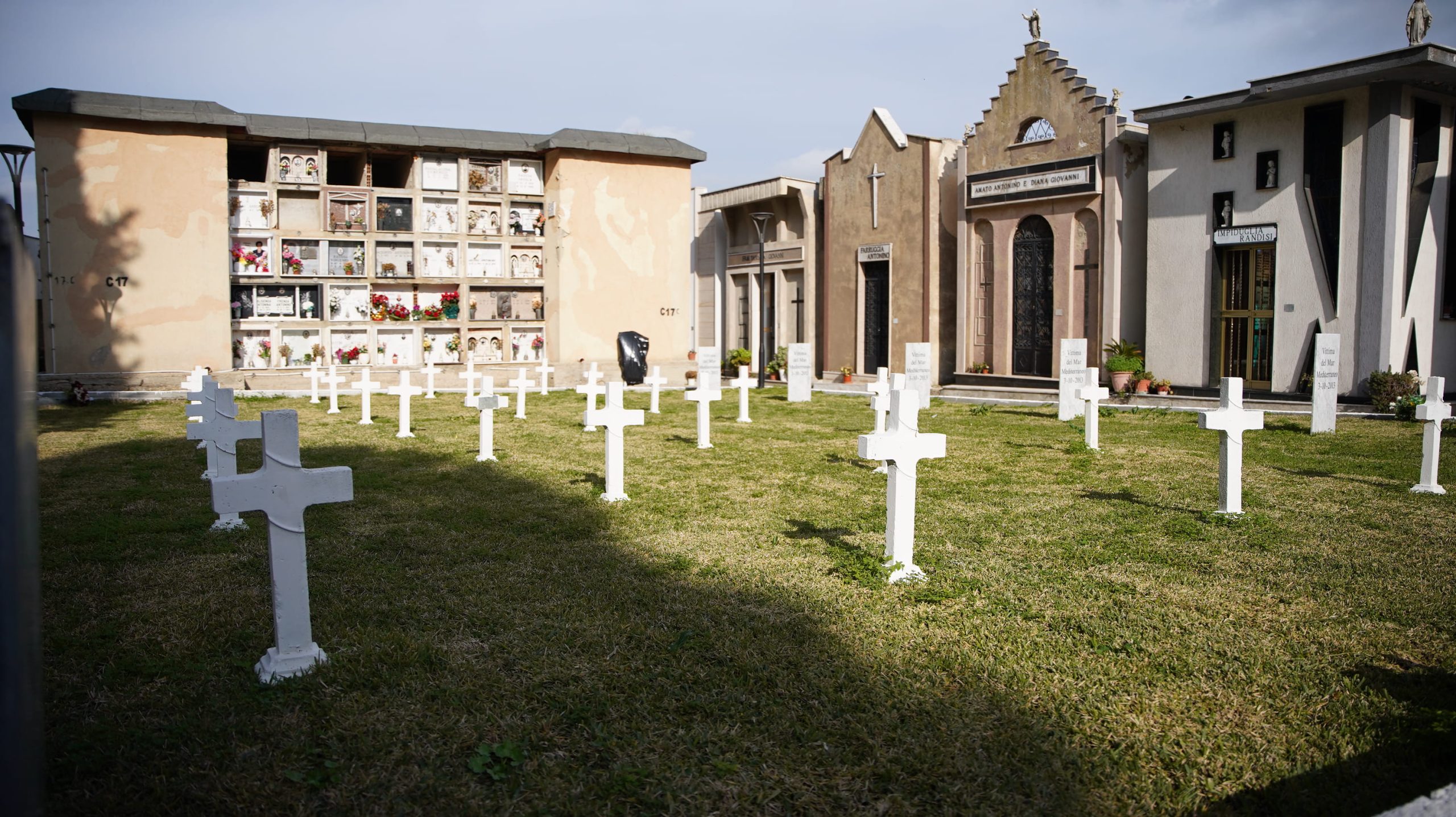 Cimitero con file di croci bianche allineate su prato verde, mausolei e cappella sullo sfondo.