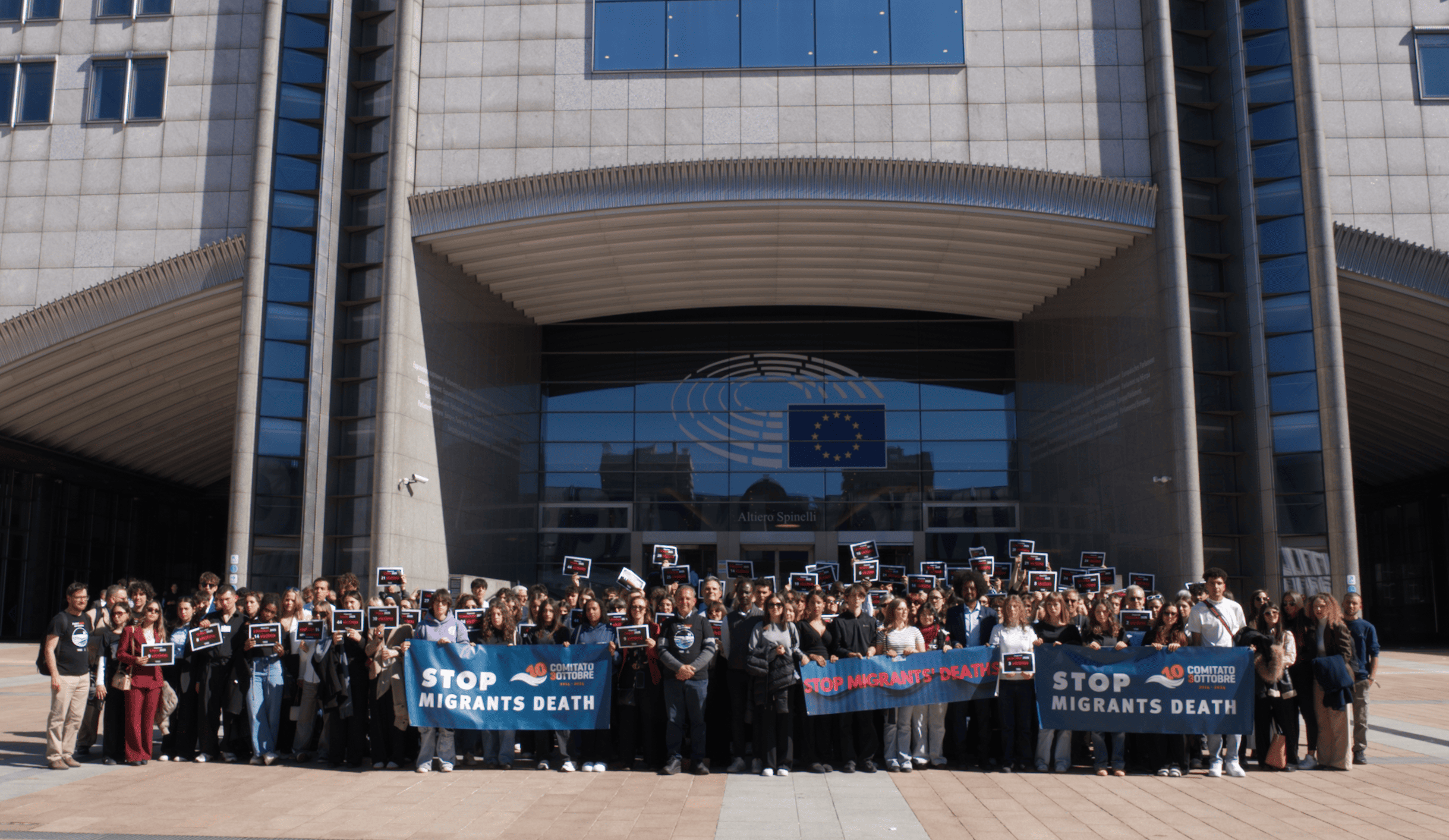 Gruppo di persone in abbigliamento informale davanti al Parlamento europeo con banner che recitano "STOP MIGRANTS DEATH".